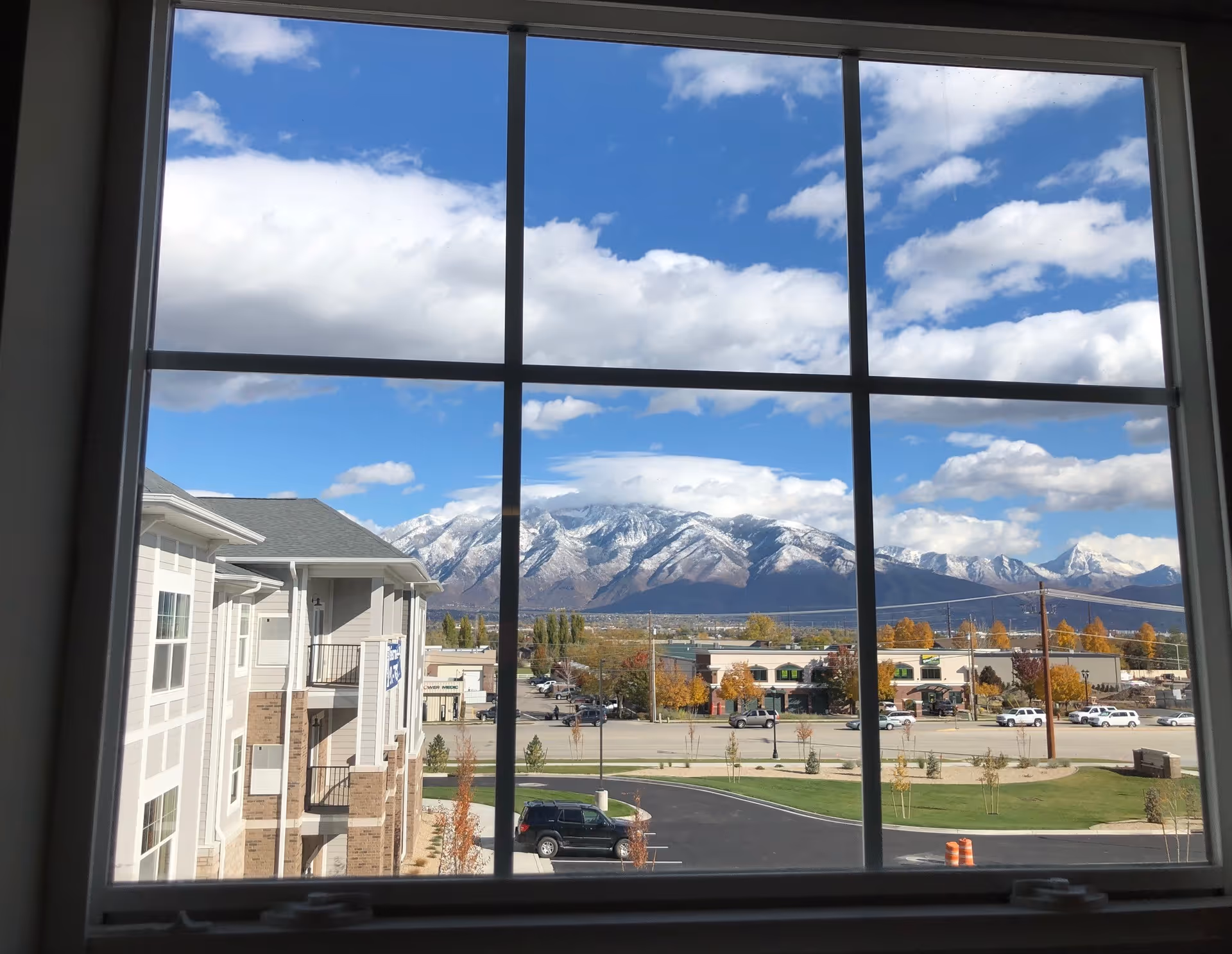View through a large window showing a parking lot, buildings, and snow-capped mountains under a partly cloudy blue sky.