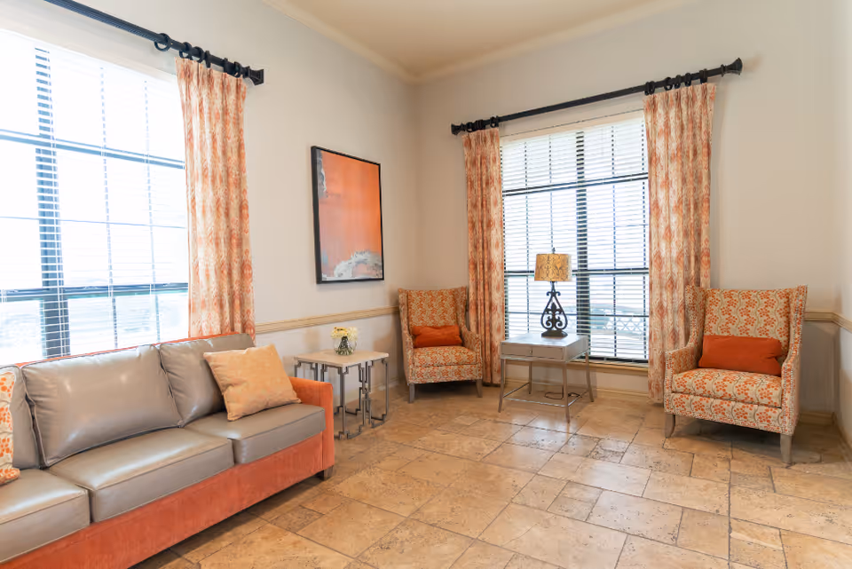 Bright sitting area with a sofa, two patterned armchairs, side tables, a lamp, and large windows with curtains.