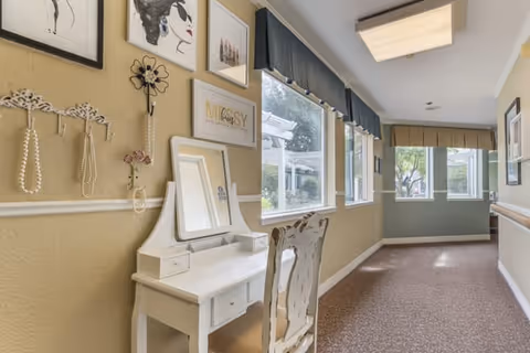 A bright hallway with beige walls and carpeted floor featuring a white vanity table and chair on the left side. The wall above the vanity is decorated with framed artwork and hanging jewelry. Large windows with blue valances let in natural light, and the hallway extends towards a green accent wall with more windows at the end.