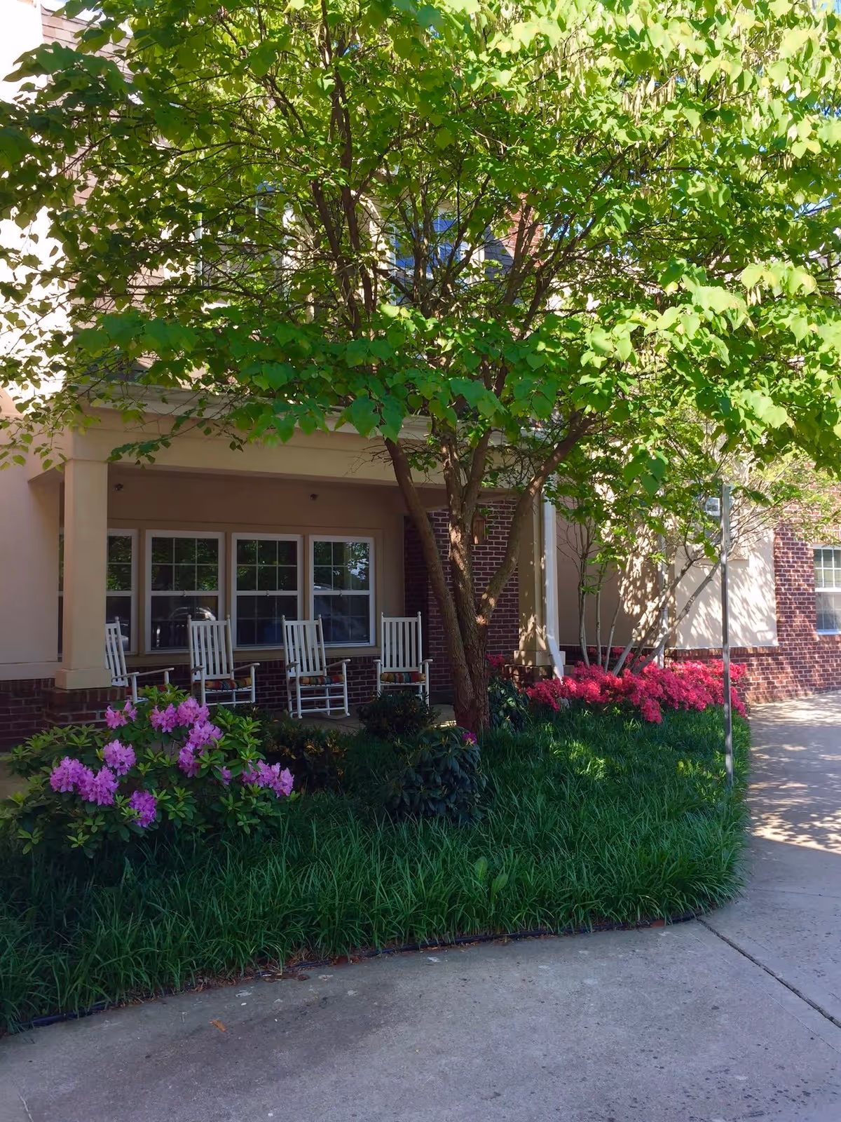 Front entrance porch with white rocking chairs beneath a large tree and landscaped flower beds along a walkway.