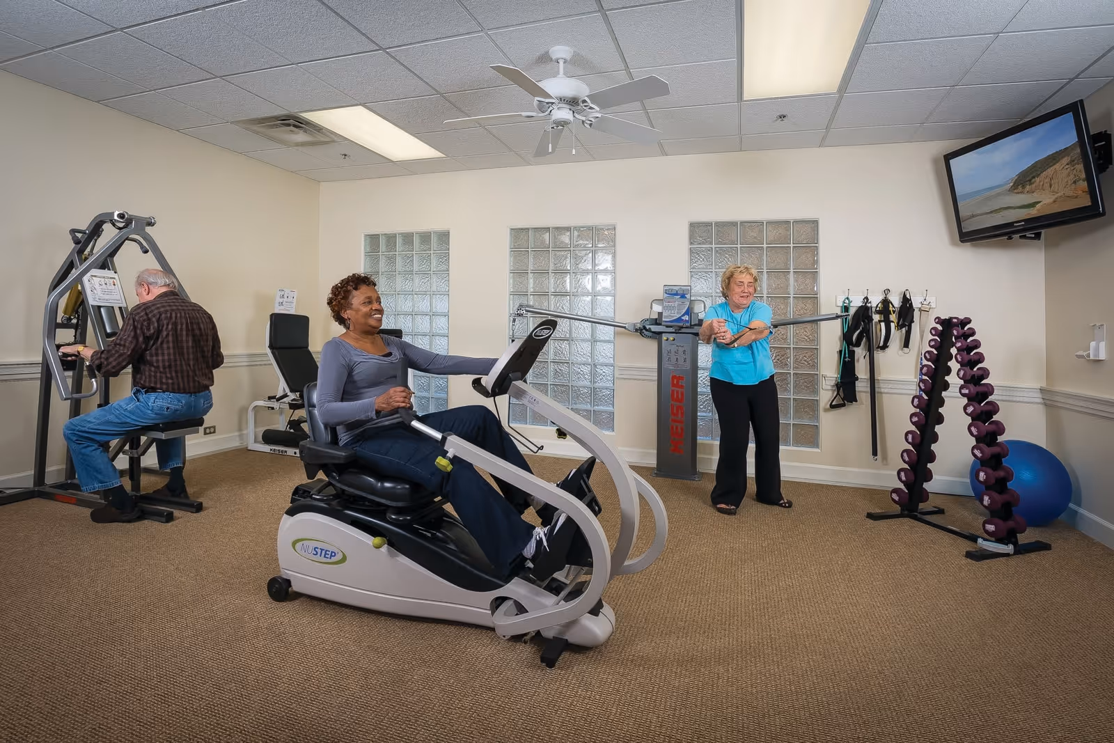 Three seniors exercising in a fitness room with various exercise equipment. One man is using a seated weight machine, a woman is on a recumbent exercise bike, and another woman is using resistance bands attached to a wall-mounted machine. A TV is mounted on the wall showing a beach scene, and a rack of dumbbells and an exercise ball are visible in the corner.