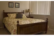 A bedroom with a wooden bed frame featuring a floral patterned bedspread and pillows. There is a wooden nightstand with a lamp next to the bed, vertical blinds covering the window, and two framed pictures hanging on the wall above the bed.