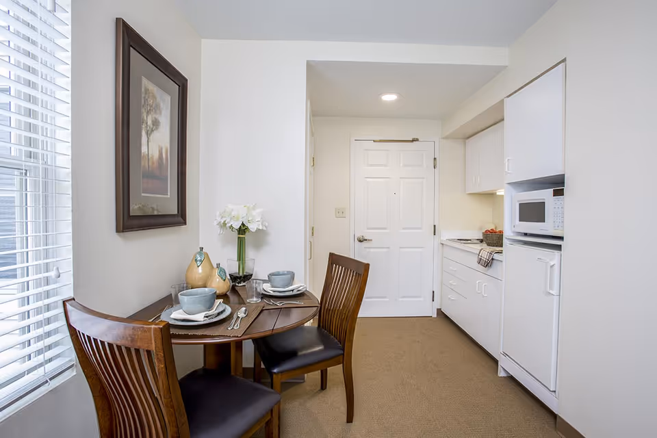 Small dining area with a round wooden table set for two with plates, bowls, glasses, and silverware. Two wooden chairs with black cushions are positioned at the table. A vase with white flowers and decorative gourds are on the table. To the right is a compact kitchen area with white cabinets, a microwave, and a small refrigerator. A white door is visible at the end of the room, and a framed picture hangs on the wall near a window with white blinds.