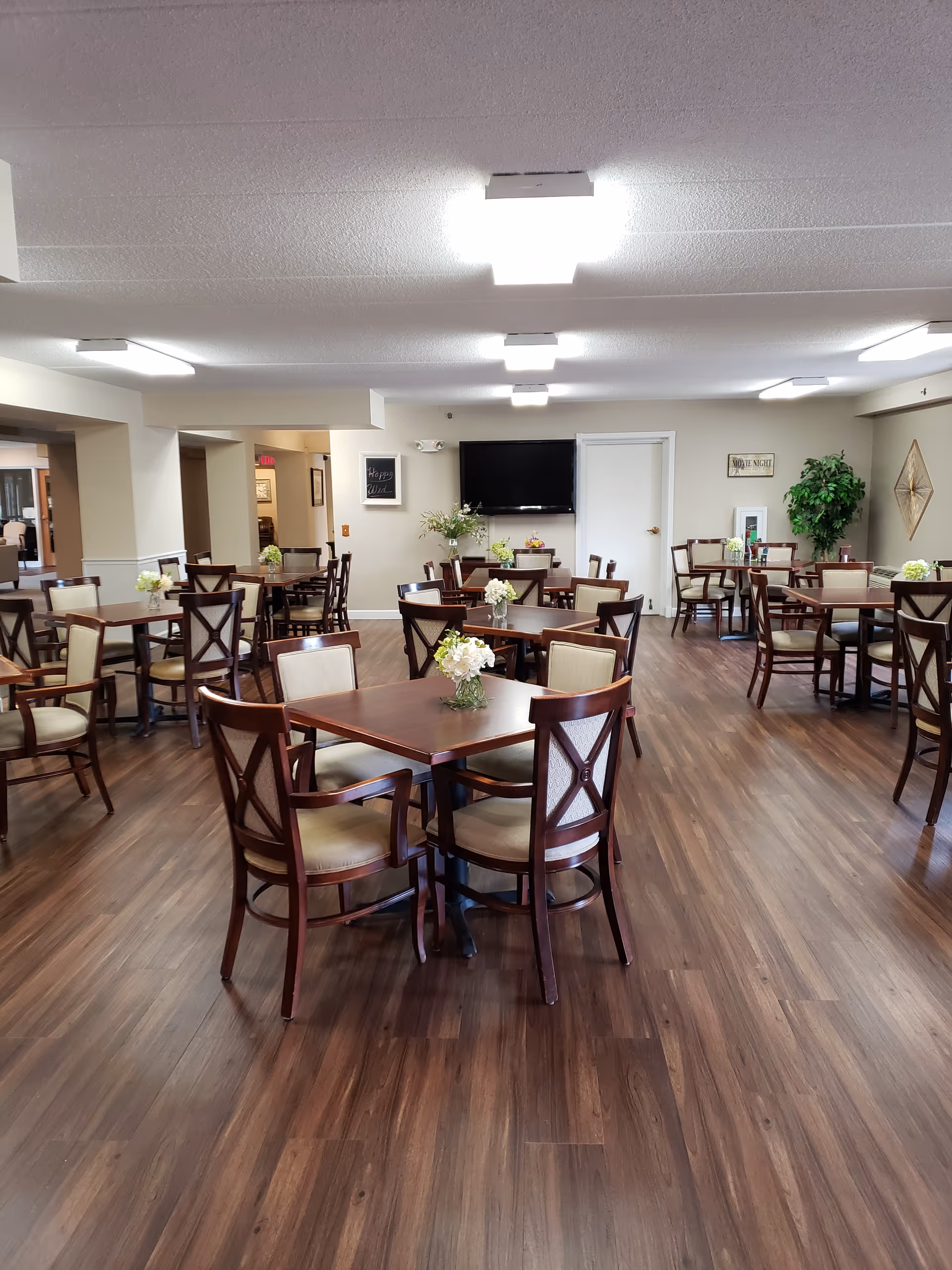 Spacious communal dining room with multiple wooden tables and chairs, floral centerpieces, and a TV on the far wall.