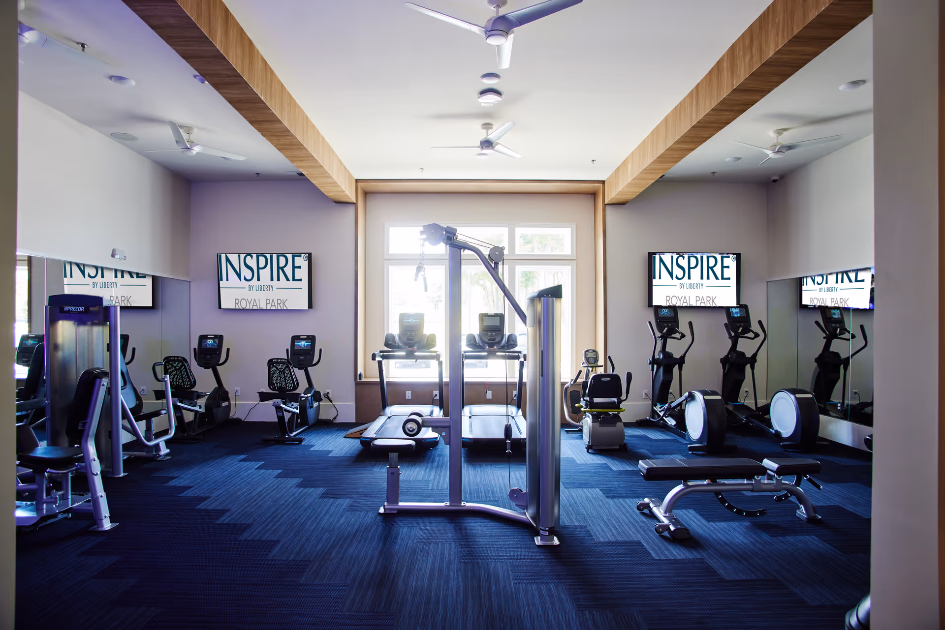 A modern fitness room with various exercise equipment including treadmills, stationary bikes, weight machines, and benches. The room has large windows letting in natural light, mirrors on the walls, and multiple ceiling fans. Two screens display the text 'INSPIRE BY LIBERTY ROYAL PARK'.