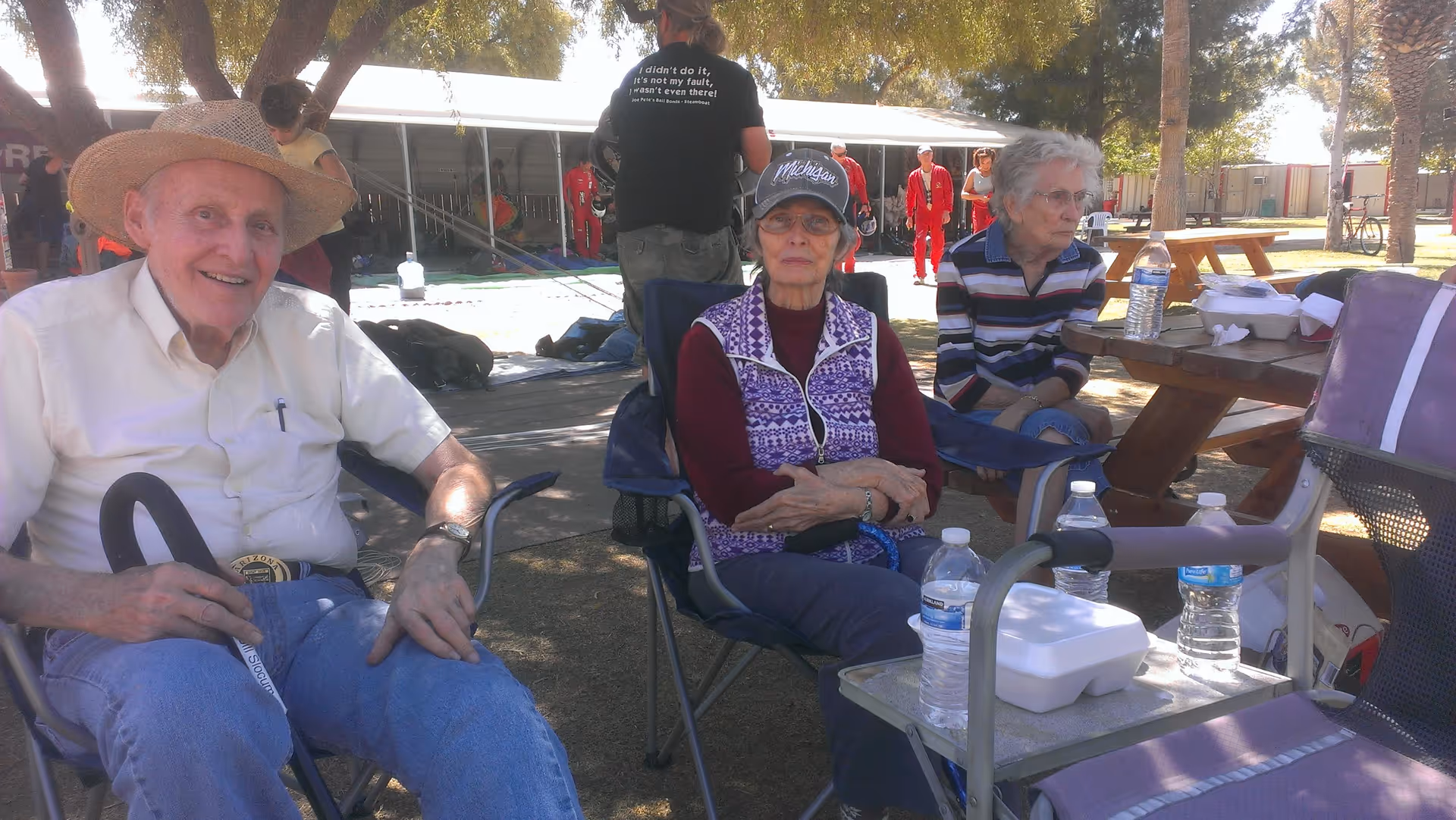 Three elderly people sitting outdoors under trees. One man wearing a straw hat and white shirt is smiling at the camera. Two elderly women are seated nearby, one wearing a Michigan cap and purple vest, the other in a striped shirt. There are picnic tables, water bottles, and food containers around them, with other people and a tent structure visible in the background.