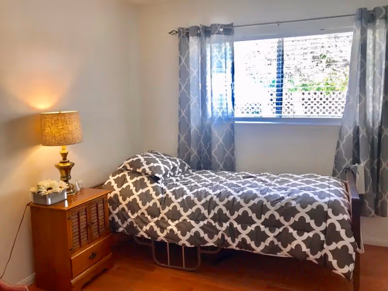 A small bedroom with a single bed covered in a gray and white patterned comforter. Next to the bed is a wooden nightstand with a lamp and a small flower arrangement. A window with gray patterned curtains lets in natural light.