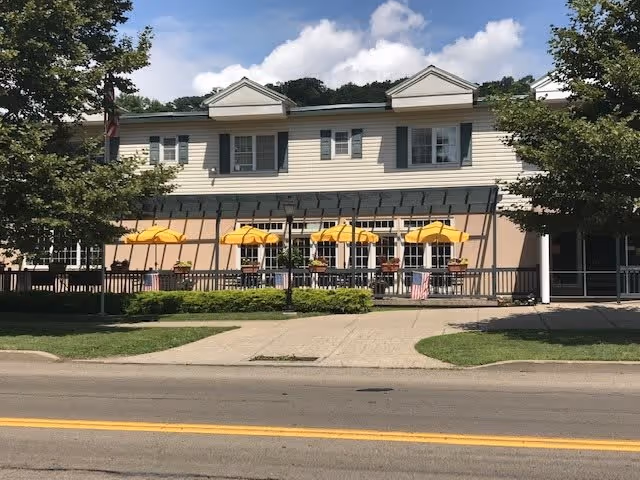 Exterior view of a two-story building with beige siding and white trim, featuring a patio area with several yellow umbrellas and outdoor seating. There are trees on either side of the building and a sidewalk in front, under a partly cloudy sky.