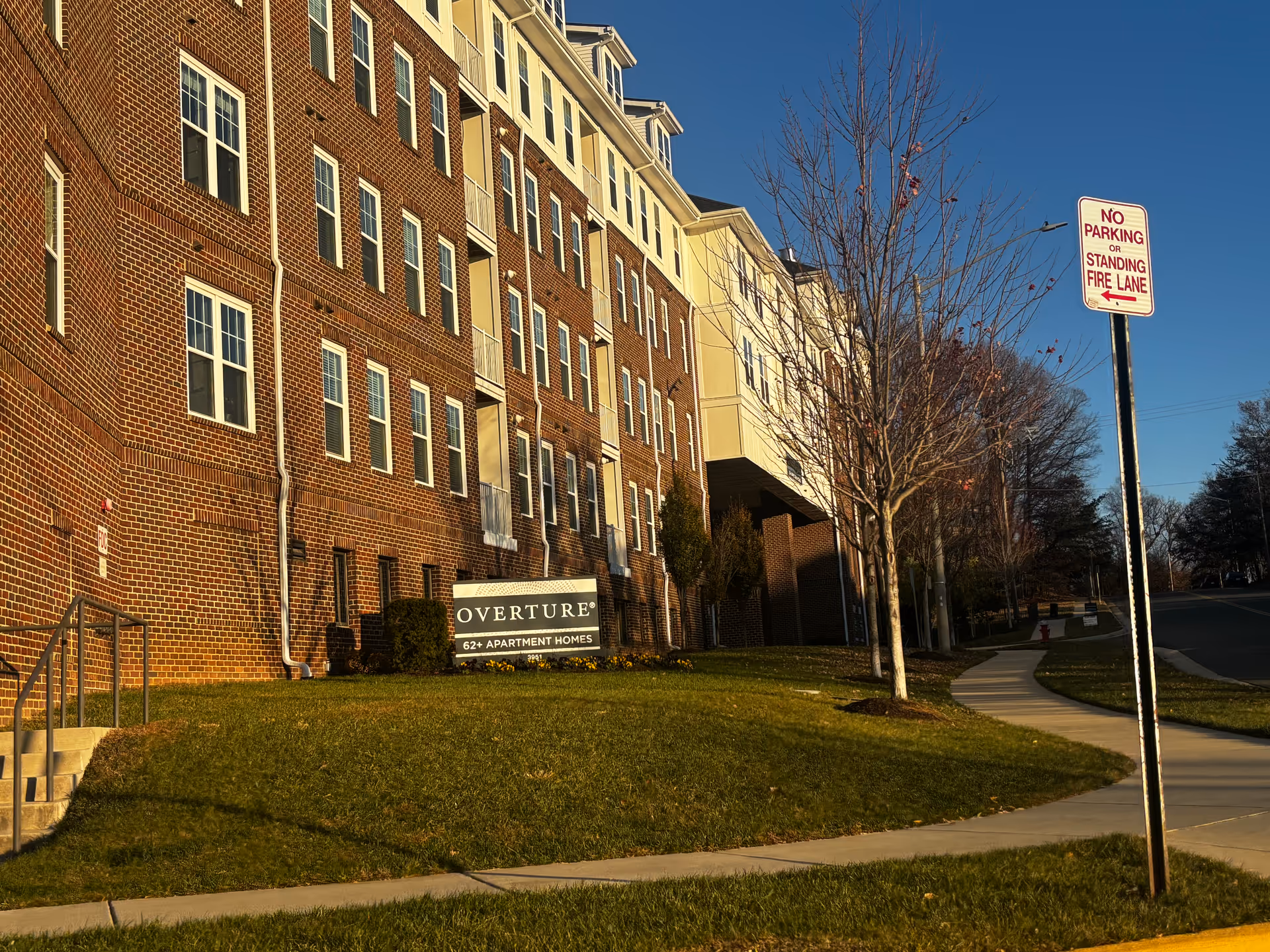 Exterior view of a multi-story brick and beige building with multiple windows and a sign in front that reads 'Overture 62+ Apartment Homes'. There is a sidewalk curving along the grass and a 'No Parking or Standing Fire Lane' sign on a pole near the street. The sky is clear and blue.