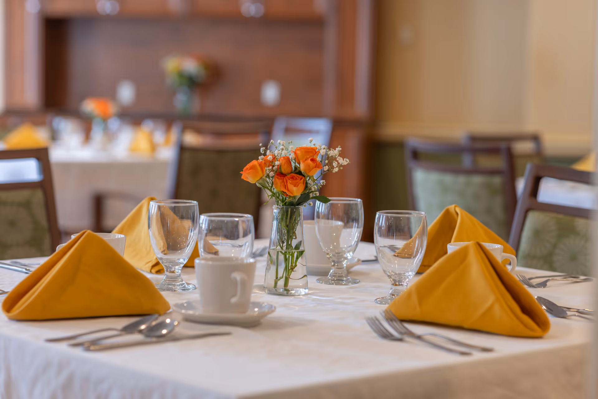 A dining table set with folded orange napkins, glassware, cutlery, a coffee cup, and a small vase of orange roses in a dining room.
