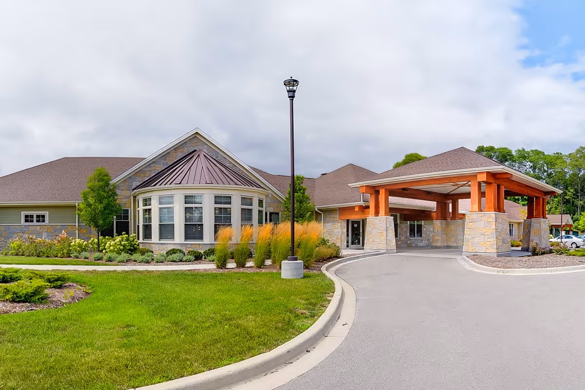 Exterior view of The Auberge at Brookfield building featuring a stone and wood facade, a covered entrance with large wooden beams, well-maintained landscaping with green grass, shrubs, and ornamental grasses, and a cloudy sky overhead.