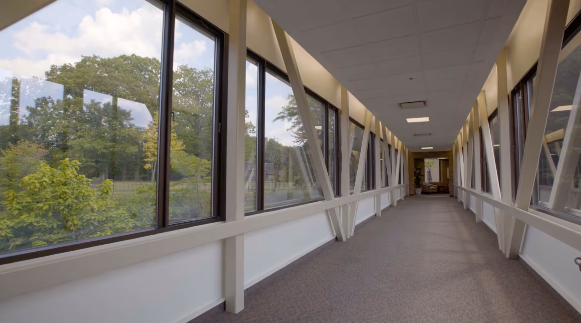 A long indoor hallway with large windows on both sides showing green trees and a partly cloudy sky outside. The hallway has a carpeted floor, white walls, and structural beams supporting the windows. At the end of the hallway, there is a seating area with chairs and a plant.