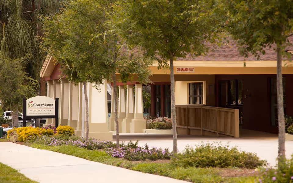 Exterior view of Grace Manor at Lake Morton, showing the entrance area with a covered driveway, surrounded by trees and landscaped bushes and flowers.