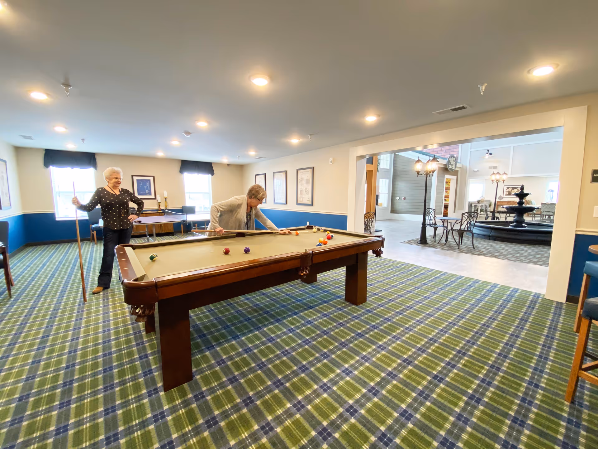 Two elderly women playing pool in a recreational room with a green and blue plaid carpet. The room has beige walls with blue wainscoting and several framed pictures. In the background, there is a ping pong table and an open area with tables, chairs, and a black fountain.