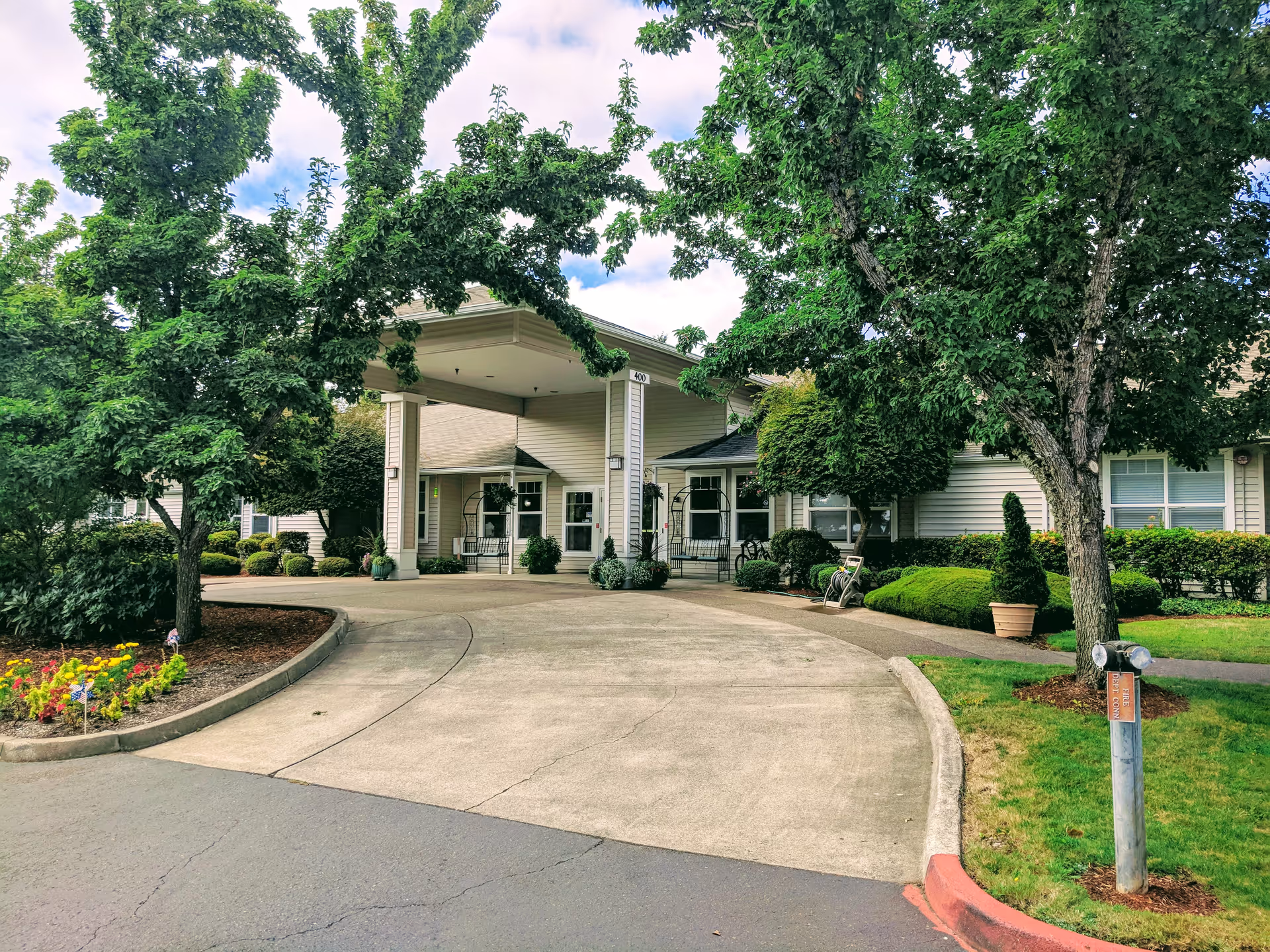Entrance of Regent Court - Assisted Living & Senior Care Community Corvallis with a covered driveway, surrounded by green trees, shrubs, and a flower bed on the left side.