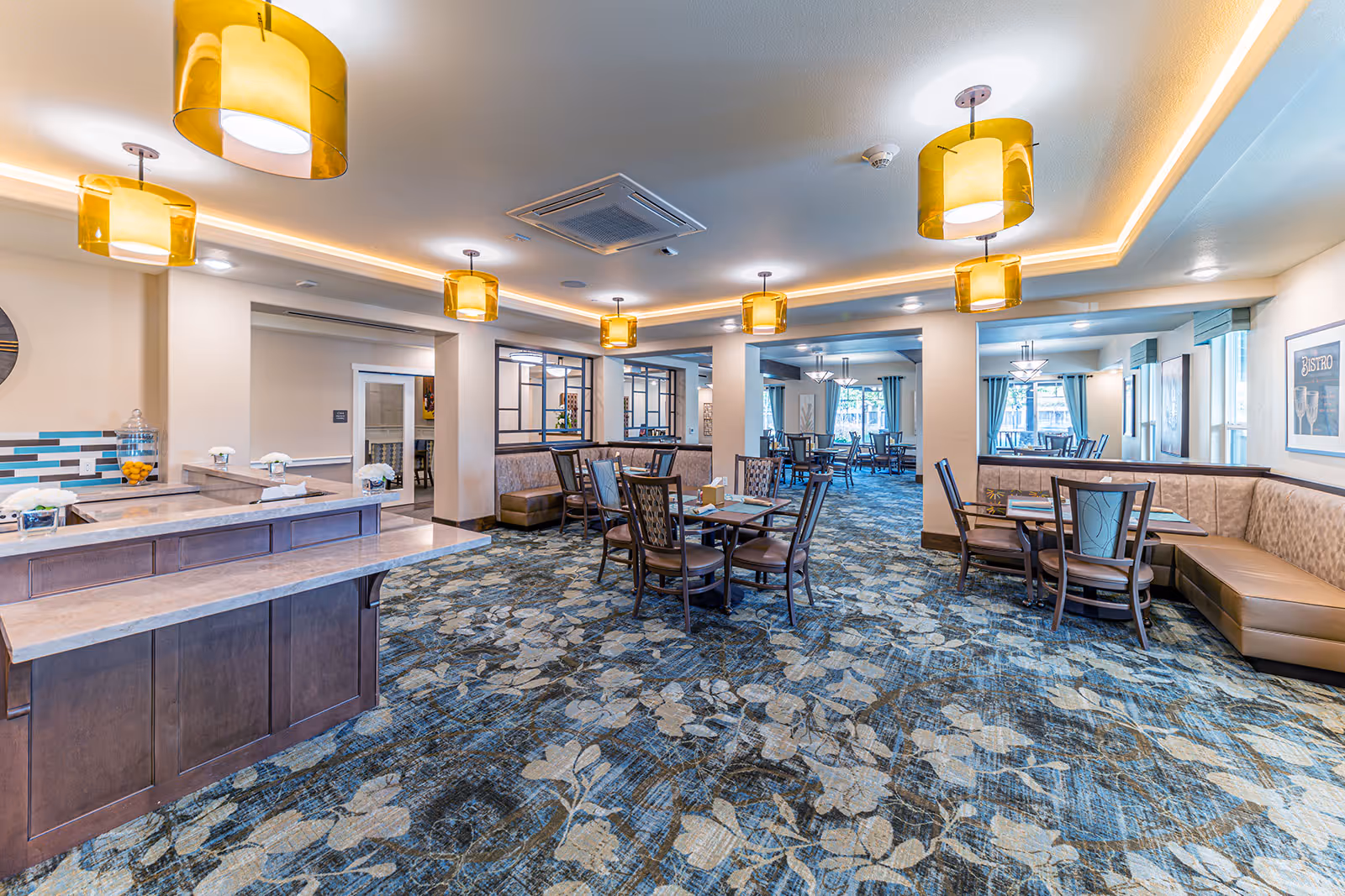 A spacious senior living dining room with multiple tables and chairs arranged on a patterned carpet. The room features warm lighting from modern pendant lamps with yellow shades, beige walls, and large windows with blue curtains allowing natural light. There is a counter area on the left side with a tiled backsplash and decorative items.