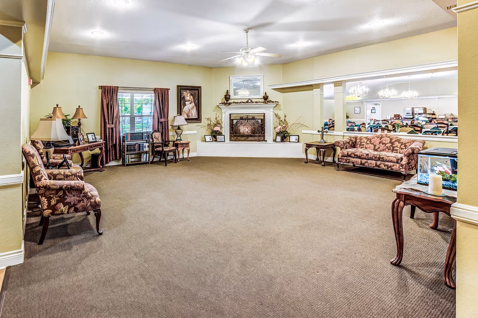 A spacious living room area in an assisted living facility featuring a carpeted floor, floral upholstered chairs and sofa, a white fireplace with decorative items on the mantel, framed artwork on the walls, and a large window with curtains. A small table with a fish tank and candle is visible in the foreground, and a dining area with tables and chairs can be seen through a large interior window opening.