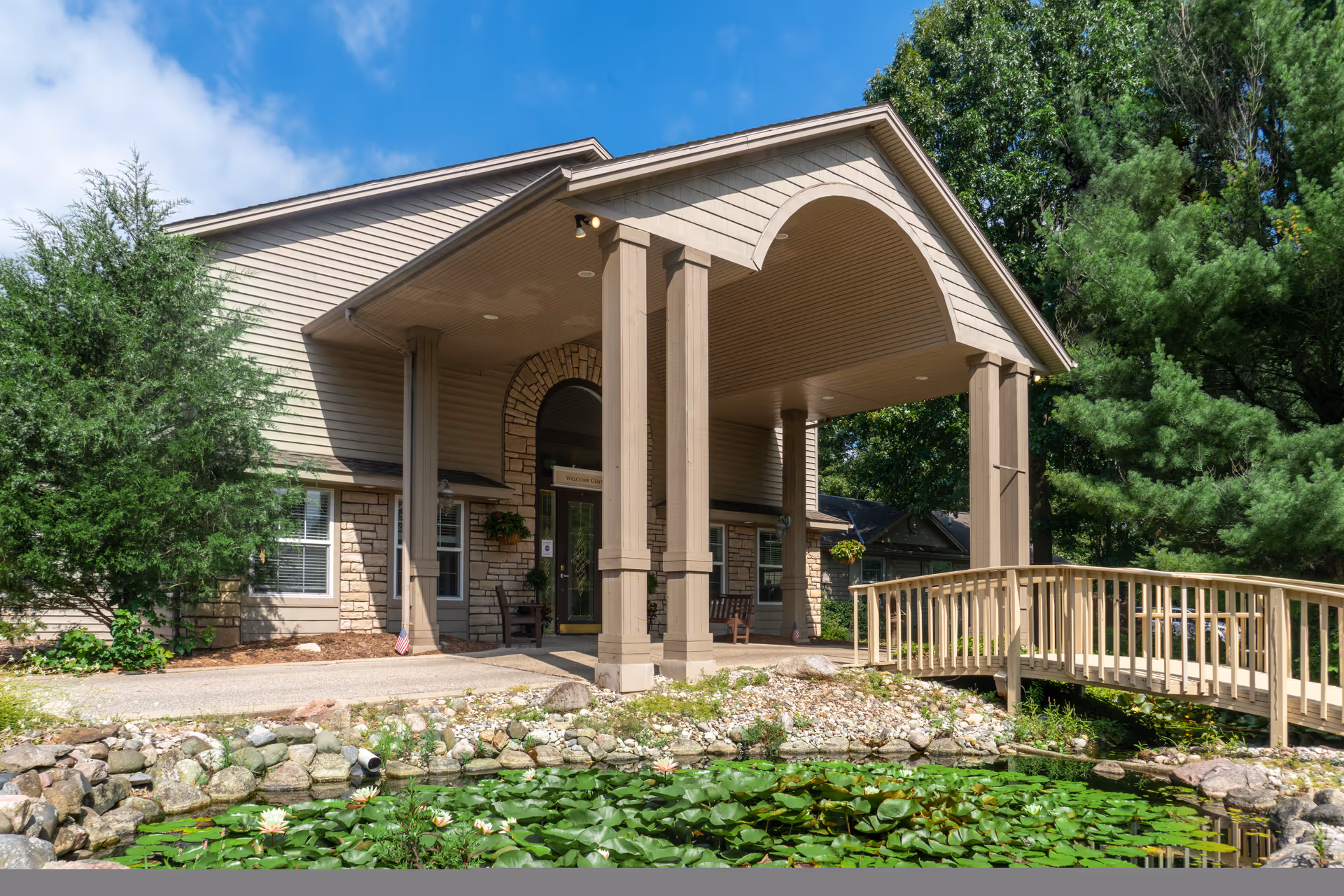 Front entrance of a senior living building with a covered portico, wooden columns, a small footbridge, and a lily-covered pond in front.