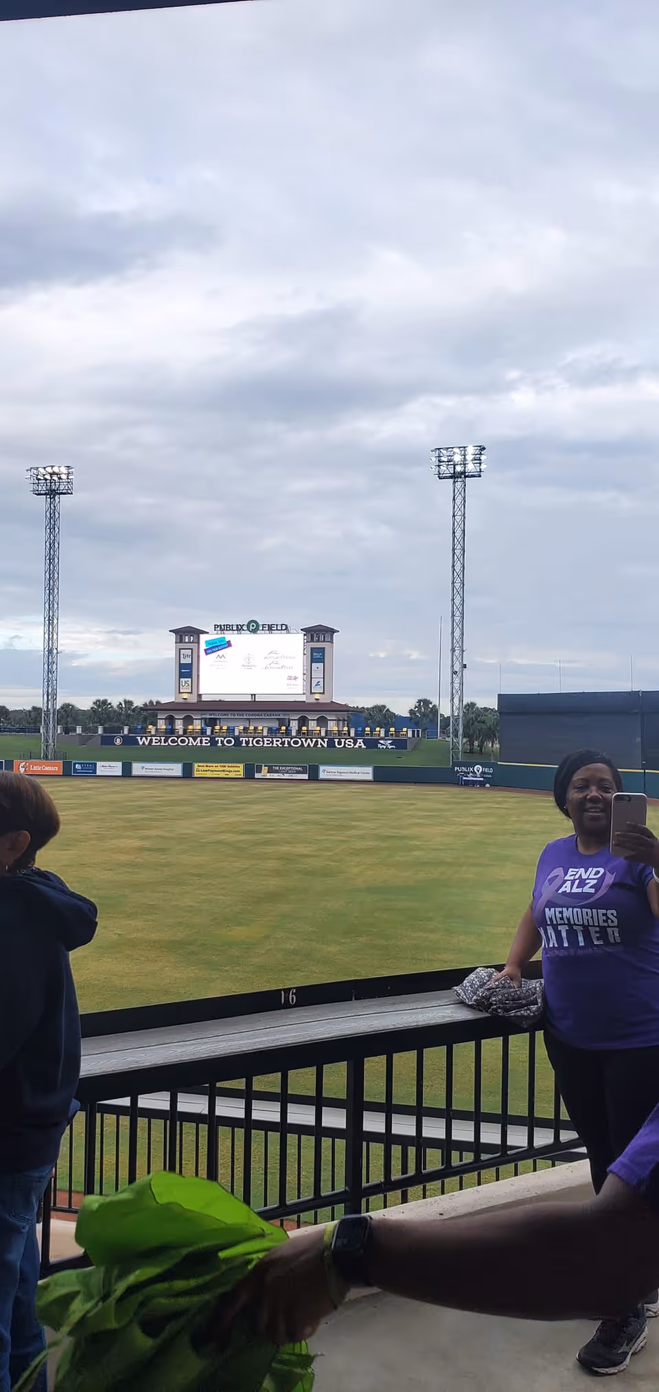 View of a baseball stadium field and scoreboard reading 'Welcome to Tigertown USA' with spectators at a railing in the foreground.