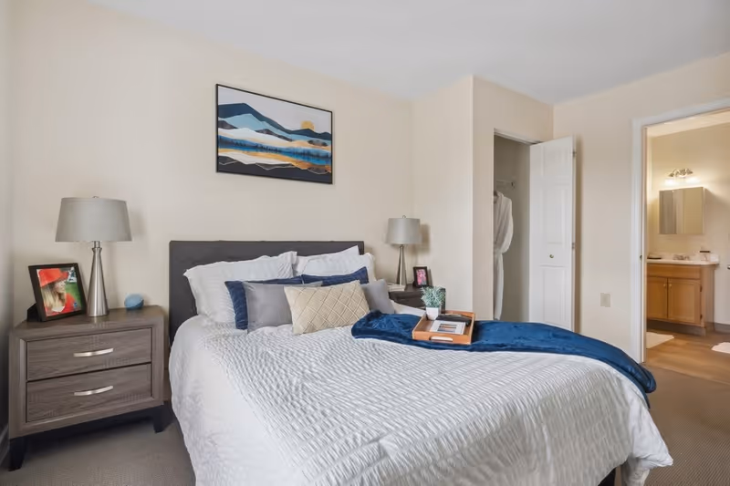 A neatly made bed with white bedding and multiple pillows in a senior living bedroom. There are two bedside tables with lamps and a framed photo on the left table. A blue throw blanket and a wooden tray with a small plant and books are on the bed. A closet with an open door and a bathroom entrance are visible in the background. A landscape painting hangs on the wall above the bed.