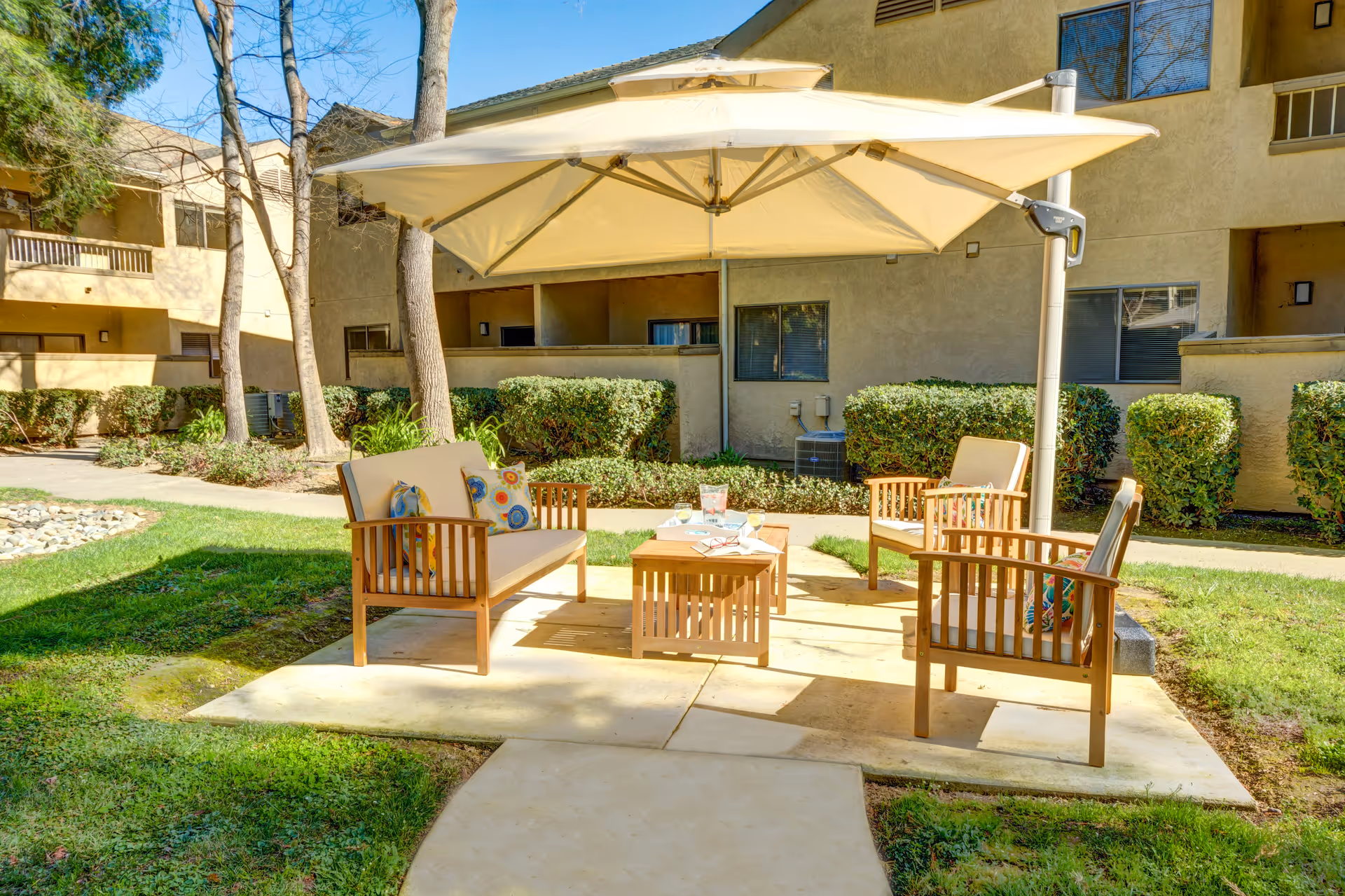 Outdoor patio area with wooden seating furniture including a loveseat and two chairs, a small wooden table with items on it, and a large beige umbrella providing shade. The patio is surrounded by grass, bushes, and trees, with a beige building in the background.