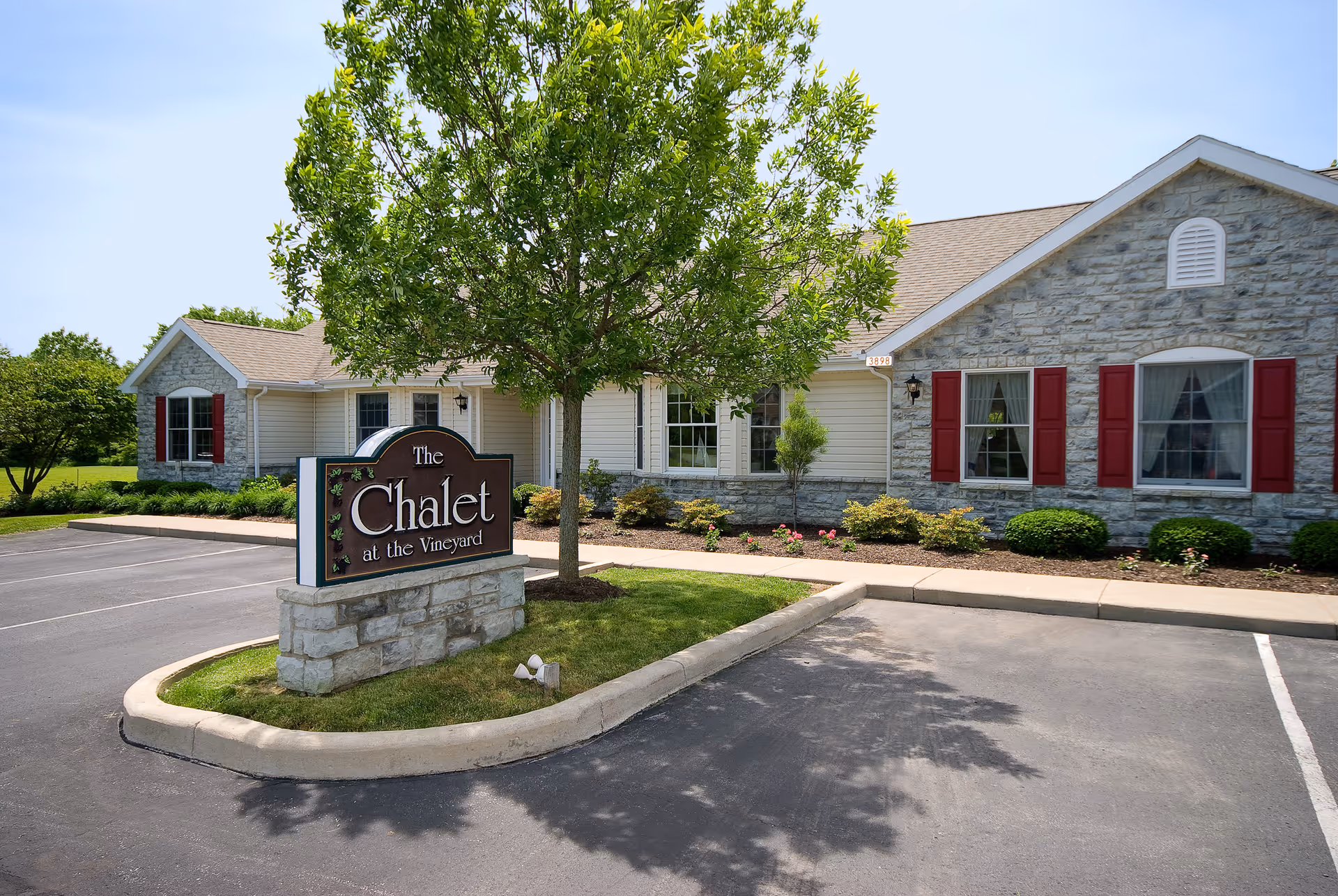 Single-story stone and siding building with red shutters and a sign reading "The Chalet at the Vineyard" beside a tree and parking lot.