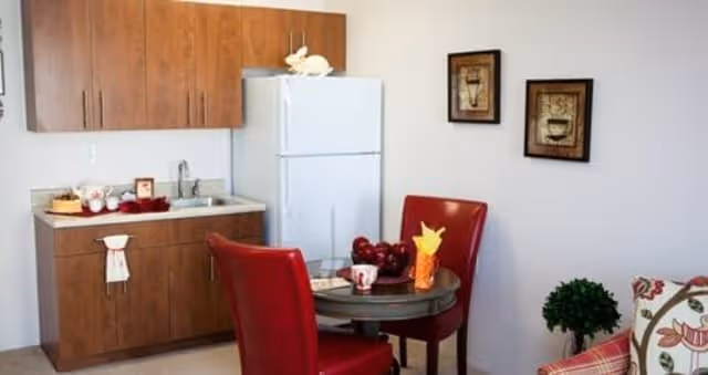A small kitchen area with wooden cabinets, a white refrigerator, and a countertop with a sink. In front of the kitchen is a round table with two red chairs. The table has a bowl of apples, a cup, and a decorative item. On the wall above the table are two framed pictures. A small green plant and part of a patterned couch are visible on the right side.
