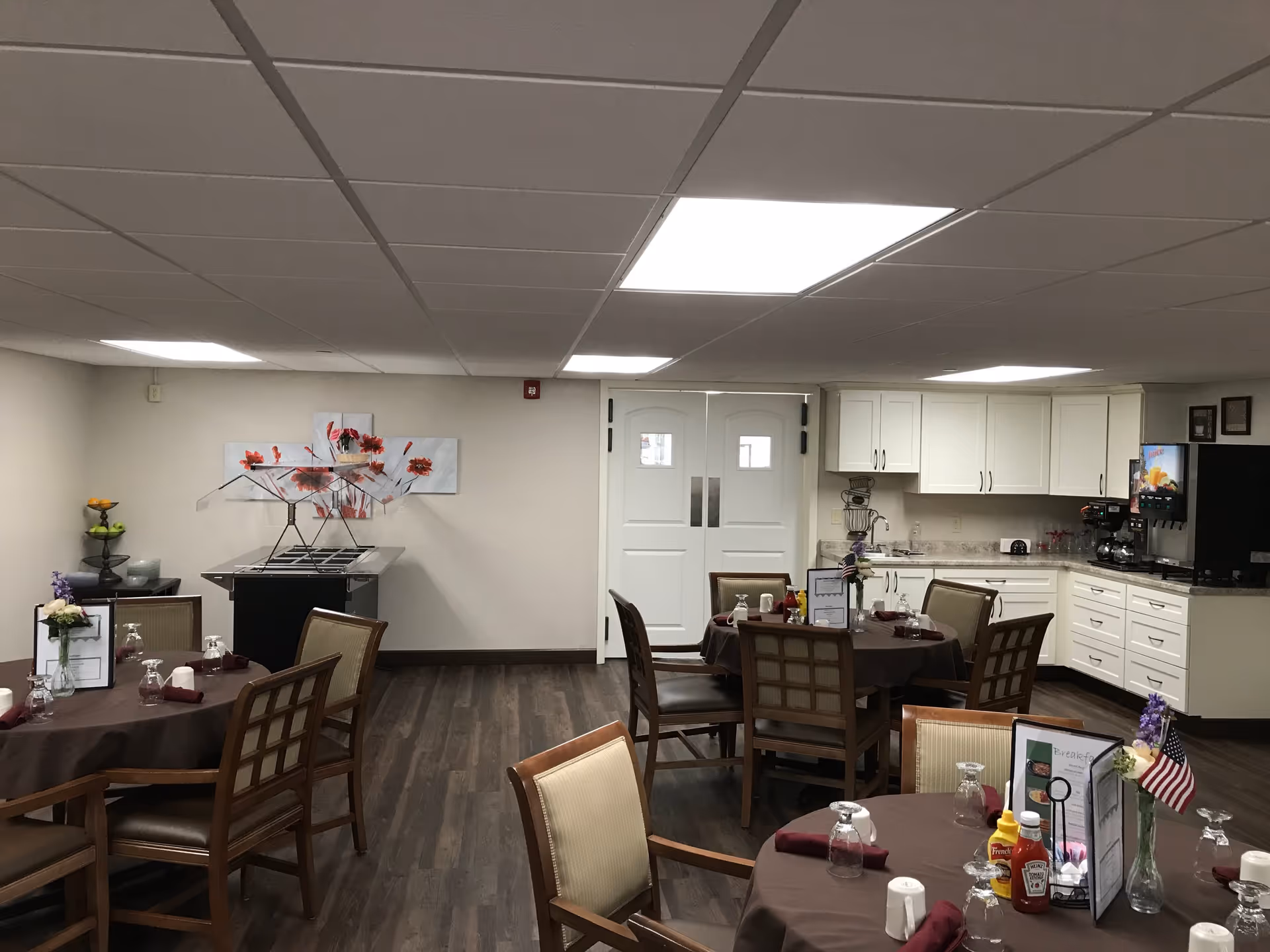 Dining area in Brown Deer Place featuring several round tables with brown tablecloths, each set with glasses, napkins, condiments, and small flower arrangements. The room has wood flooring, white walls, and a ceiling with recessed lighting. In the background, there is a white double door, a buffet station, and a kitchen area with white cabinets and a coffee machine.
