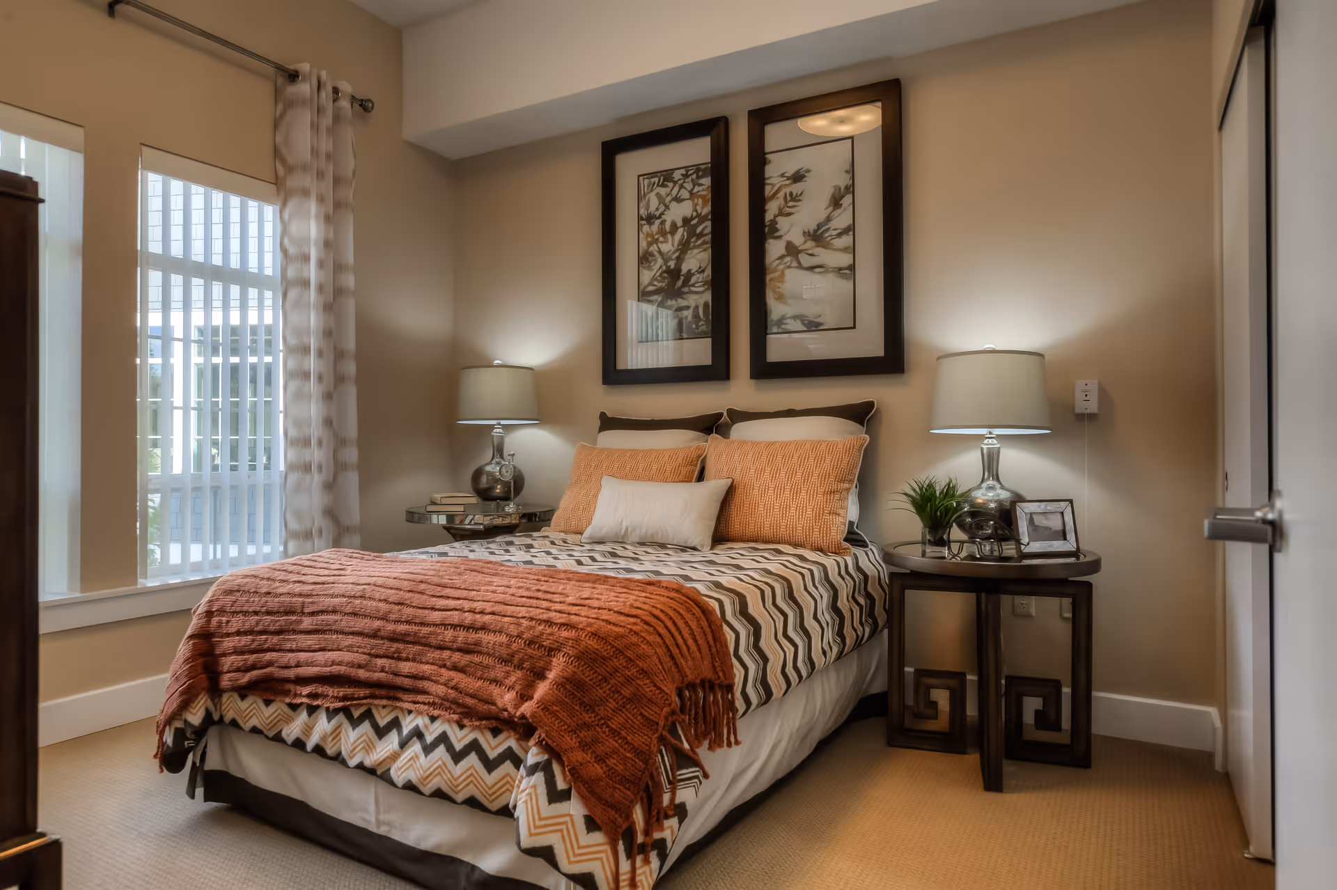 A neatly staged bedroom with a bed dressed in patterned bedding and an orange throw, two bedside tables with lamps, and framed artwork above the headboard.