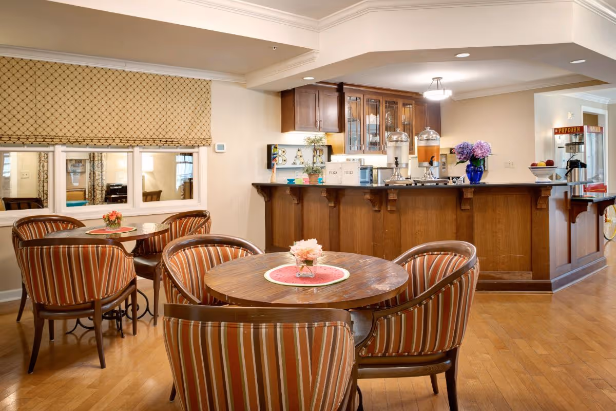 A cozy dining area with round wooden tables and striped upholstered chairs facing a wooden service bar with drink dispensers and a popcorn machine.