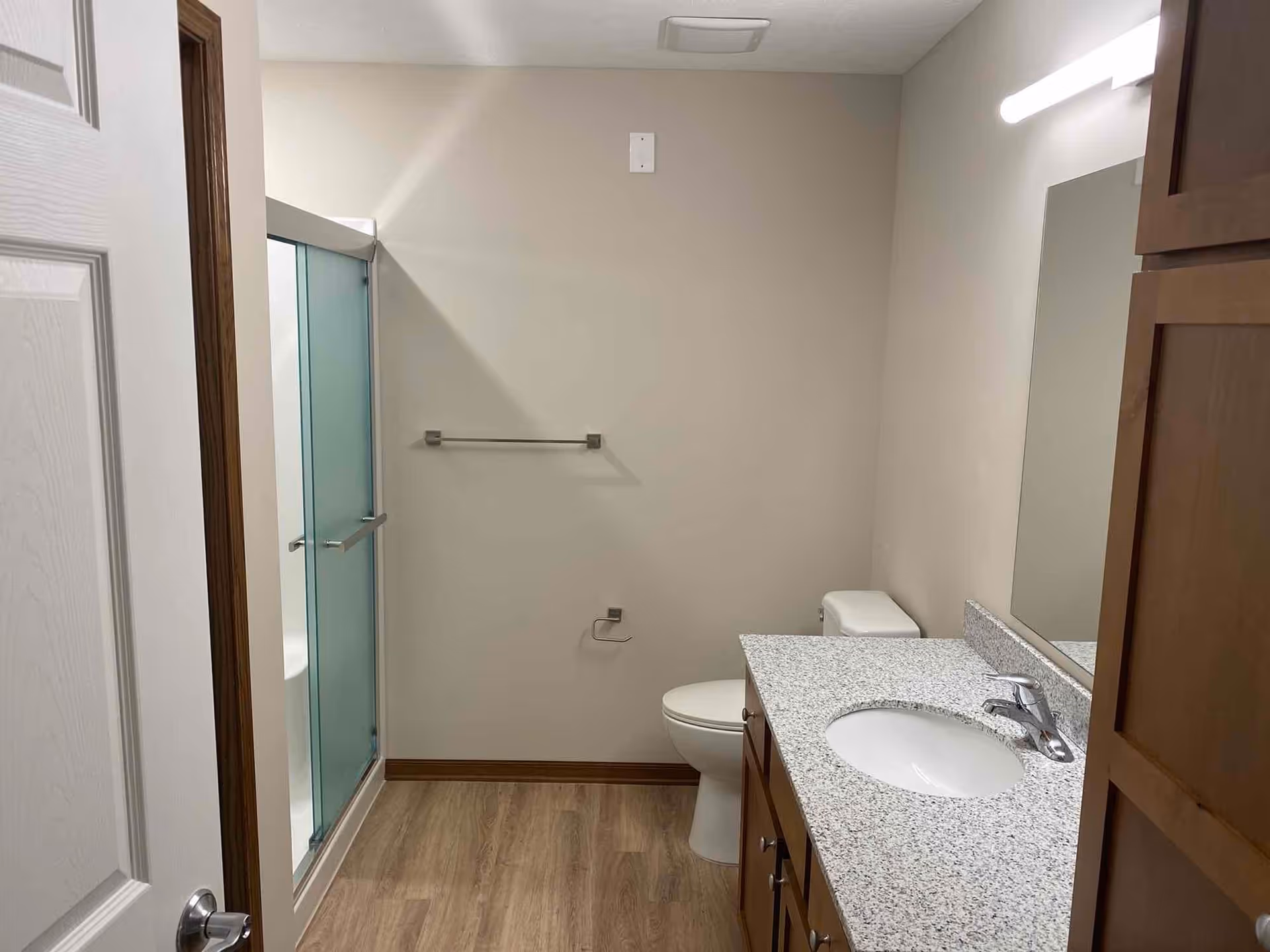 A clean bathroom with a wooden door partially open, a glass-enclosed shower on the left, a toilet next to a granite countertop with a sink, a large mirror above the sink, and wooden cabinets. The floor has a wood-like finish and the walls are painted light beige.