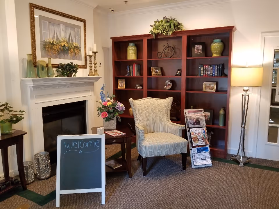 Lobby seating area with a striped armchair in front of a wooden bookshelf, a decorated fireplace mantel, flowers, a floor lamp, and a small chalkboard sign reading "welcome".