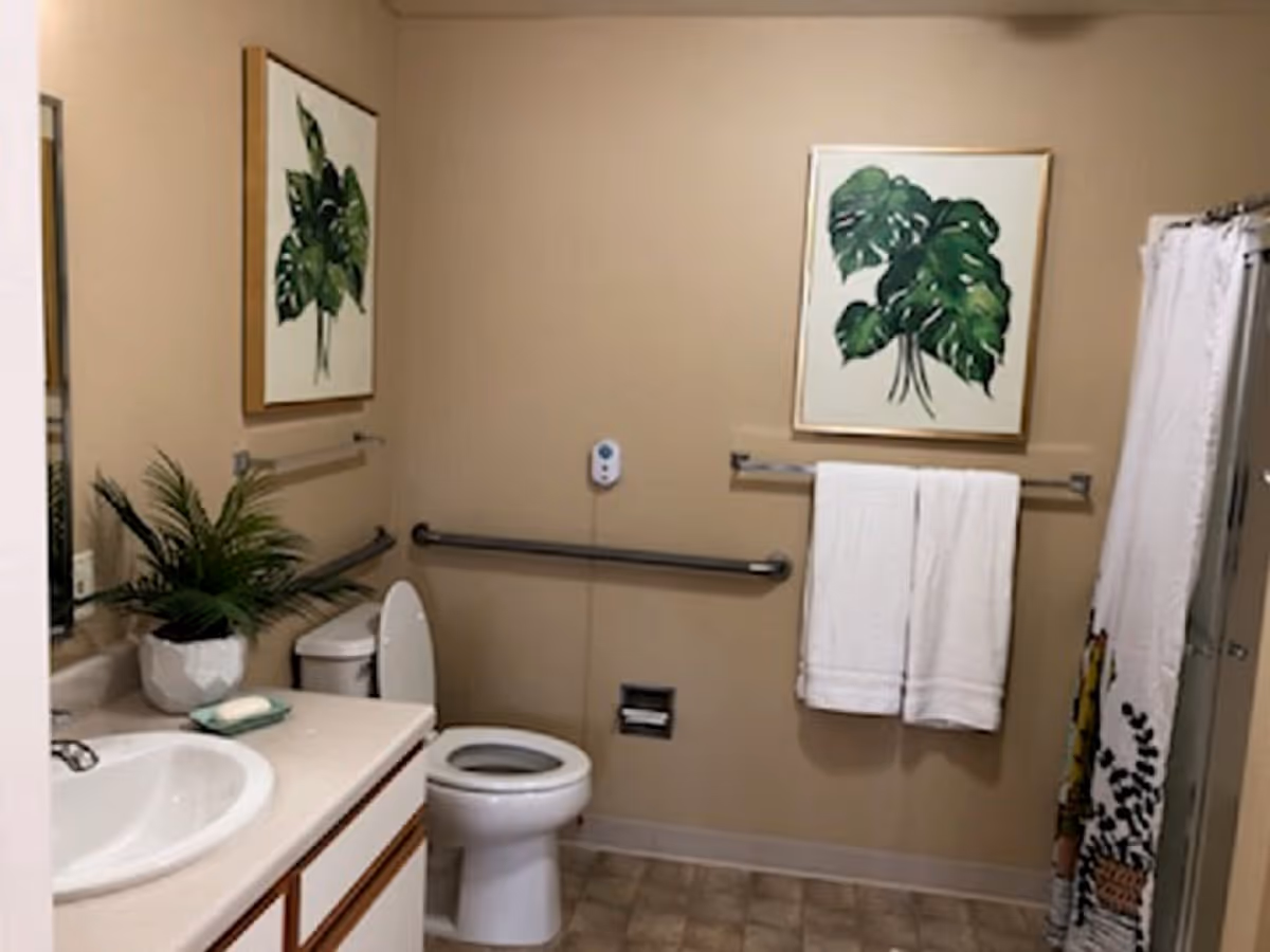 A clean bathroom with beige walls featuring a white toilet with the lid open, a white sink with a bar of soap and a potted plant on the counter, two framed botanical prints on the walls, a towel rack with two white towels, and a shower with a white curtain decorated with plant designs.