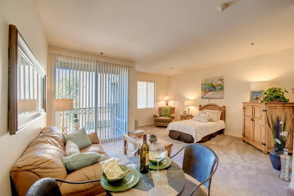 A bright and cozy senior living apartment featuring a leather sofa with cushions, a small round dining table set with plates, napkins, wine glasses, and a bottle of wine. The room includes a single bed with white bedding, a wooden nightstand with a lamp, an armchair with a green throw, a wooden cabinet, and decorative plants. Large sliding glass doors with vertical blinds lead to a balcony, allowing natural light to fill the space.