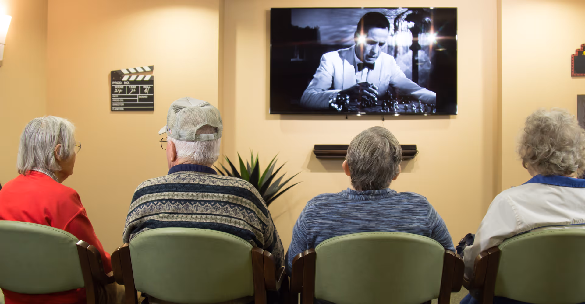 Four elderly individuals sitting in green chairs facing a wall-mounted television screen showing a black and white movie scene with a man in a suit. The room has beige walls and a small plant is visible between the chairs and the TV.