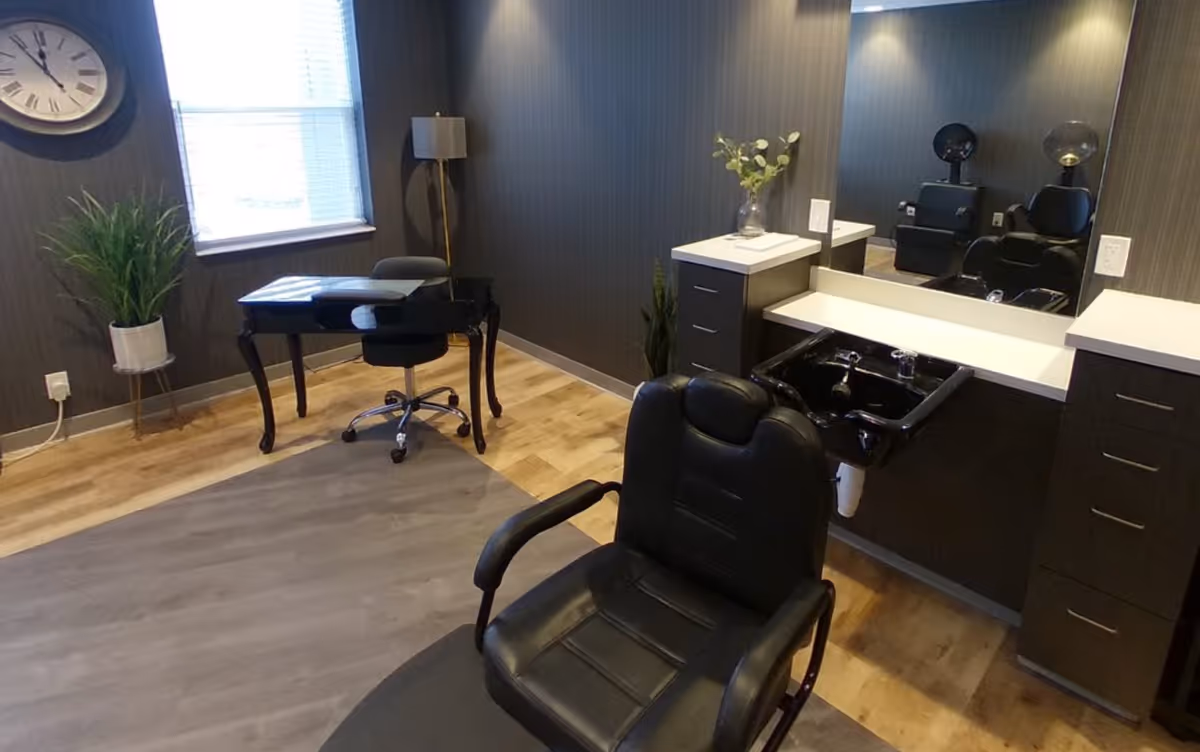 A bright interior salon room with a black styling chair in front of a sink and mirror, a desk by the window, plants and a wall clock.