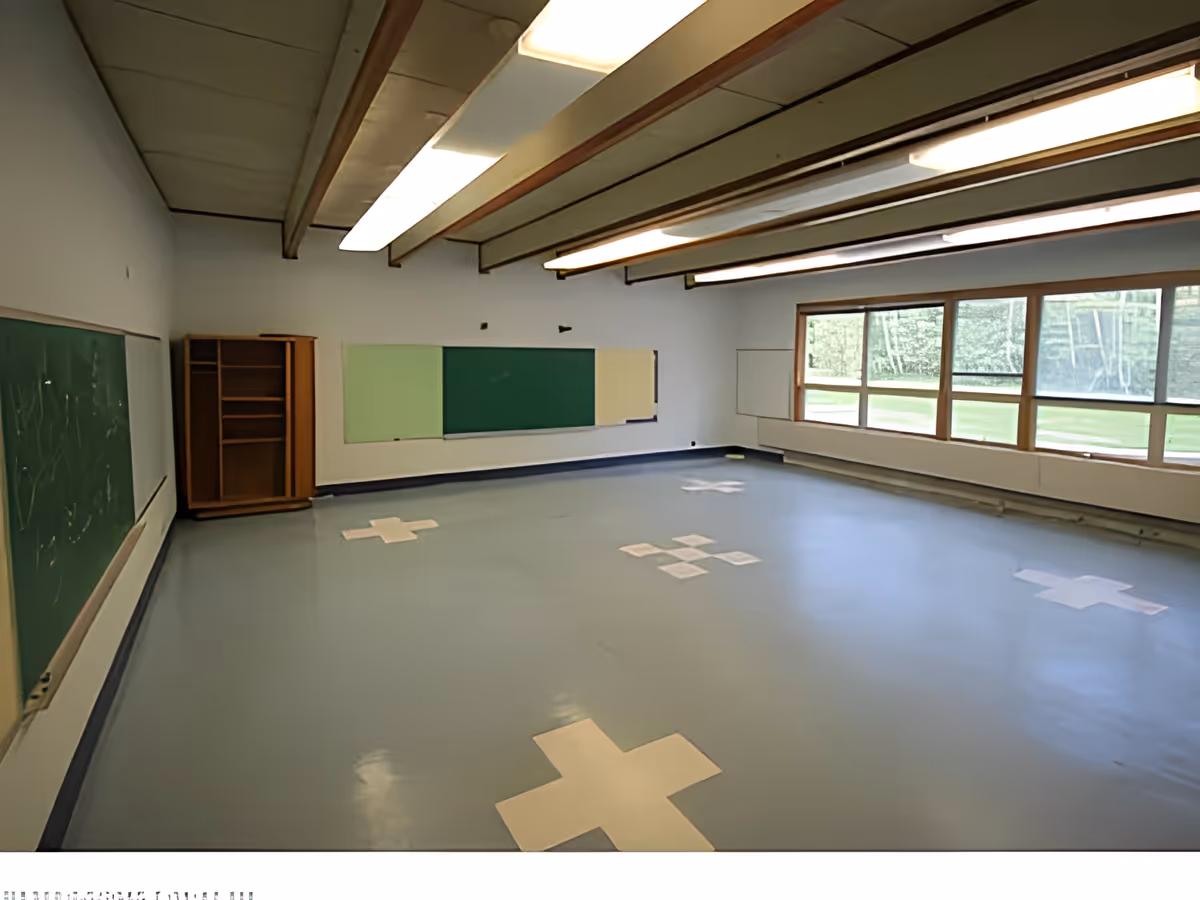 Empty multipurpose interior room with blue floor marked by white cross shapes, chalkboards on the walls, large windows and overhead fluorescent lights.