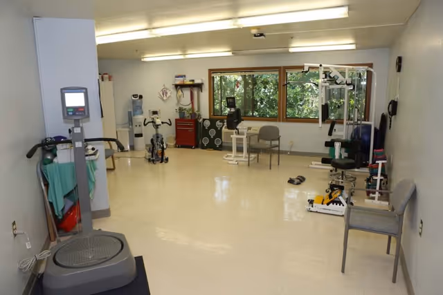A rehabilitation exercise room with various fitness equipment including stationary bikes, a vibration plate machine, resistance training machines, chairs, and a water cooler. Large windows provide natural light and a view of greenery outside.