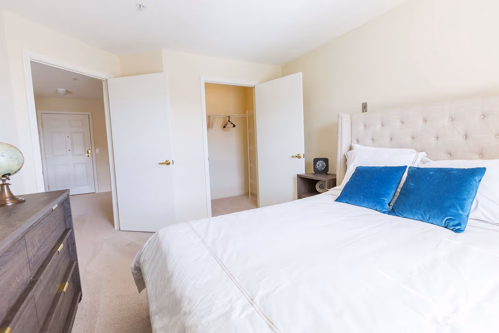 A bright bedroom with a large bed featuring a tufted beige headboard and white bedding accented with two blue pillows. To the right of the bed is a small wooden nightstand with a black clock. On the left side of the image is a wooden dresser with a globe on top. Two open doors reveal a walk-in closet with a few hangers and an entrance door in the background. The room has light-colored walls and carpeted flooring.