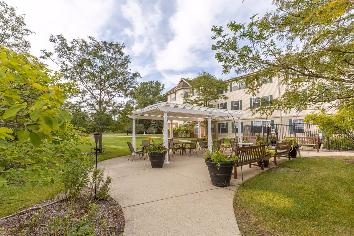 Outdoor patio area at StoryPoint Libertyville featuring a white pergola with several tables and chairs underneath. There are large potted plants around the patio, wooden benches, and a well-maintained lawn with trees and shrubs surrounding the space. The building with multiple windows is visible in the background under a partly cloudy sky.