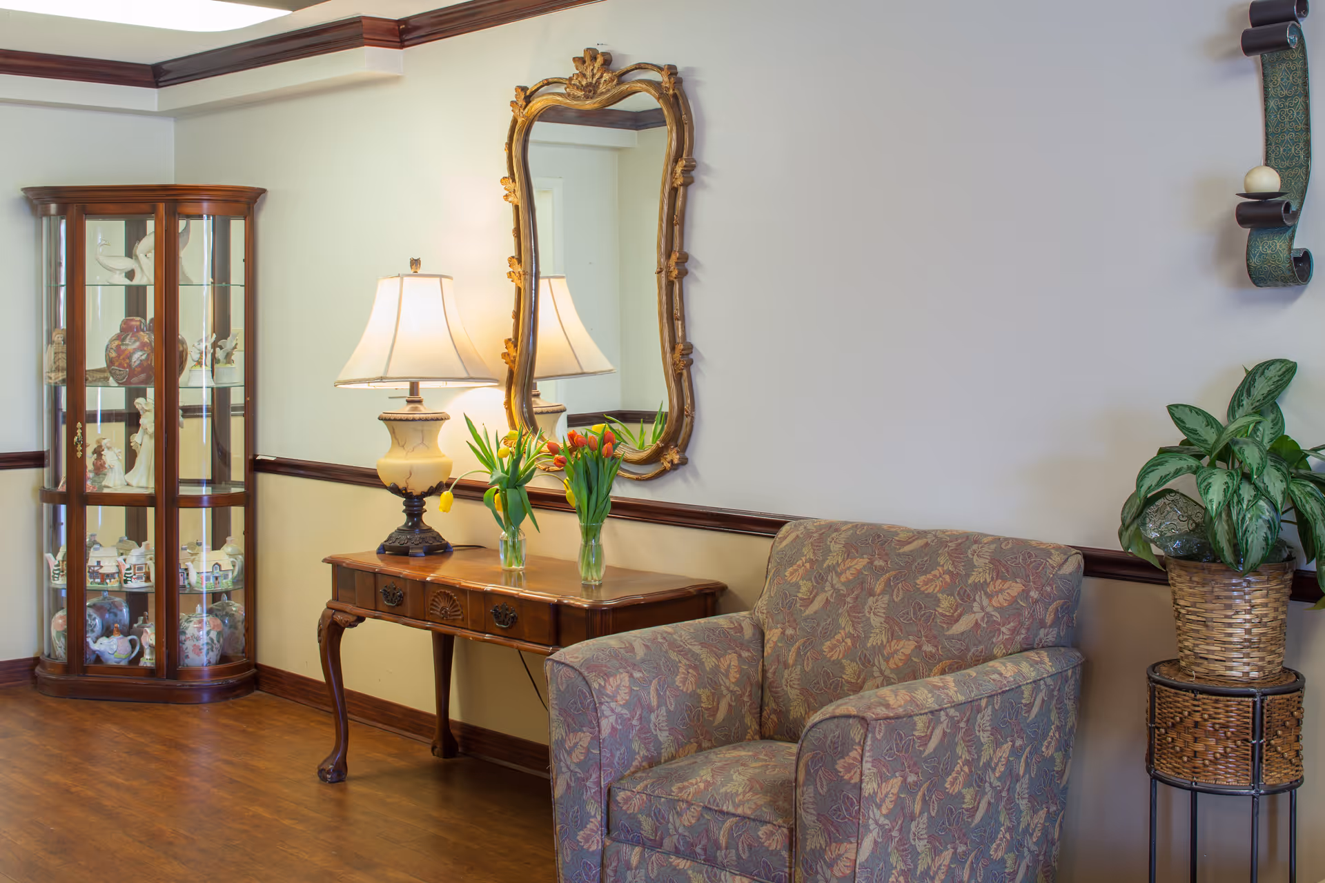 A cozy interior corner featuring a floral-patterned armchair, a wooden side table with a decorative lamp and two vases of tulips, a large ornate mirror hanging on the wall, a glass display cabinet with various decorative items, and a potted plant on a woven stand.