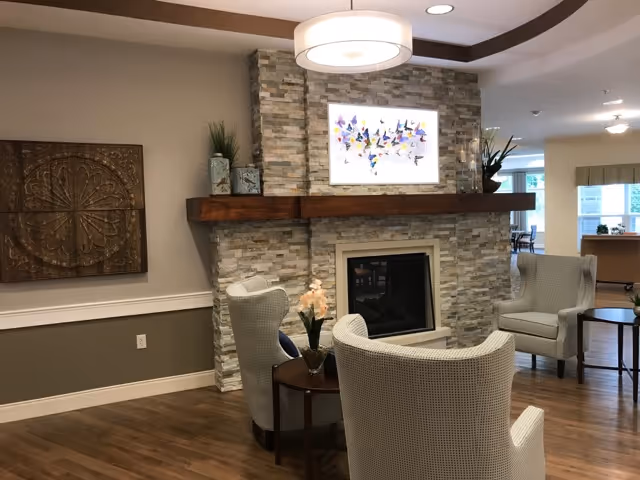 Cozy lounge area featuring a stone fireplace with wooden mantel, upholstered armchairs, side tables, and wall art.