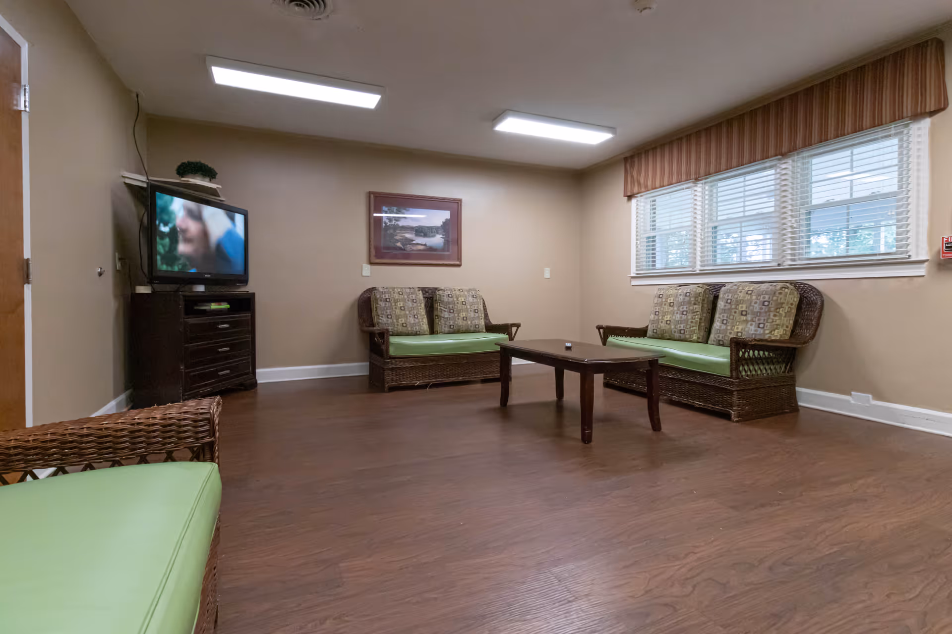 A common-area lounge with wicker sofas with green cushions, a low coffee table, a TV on a stand, and windows with blinds.