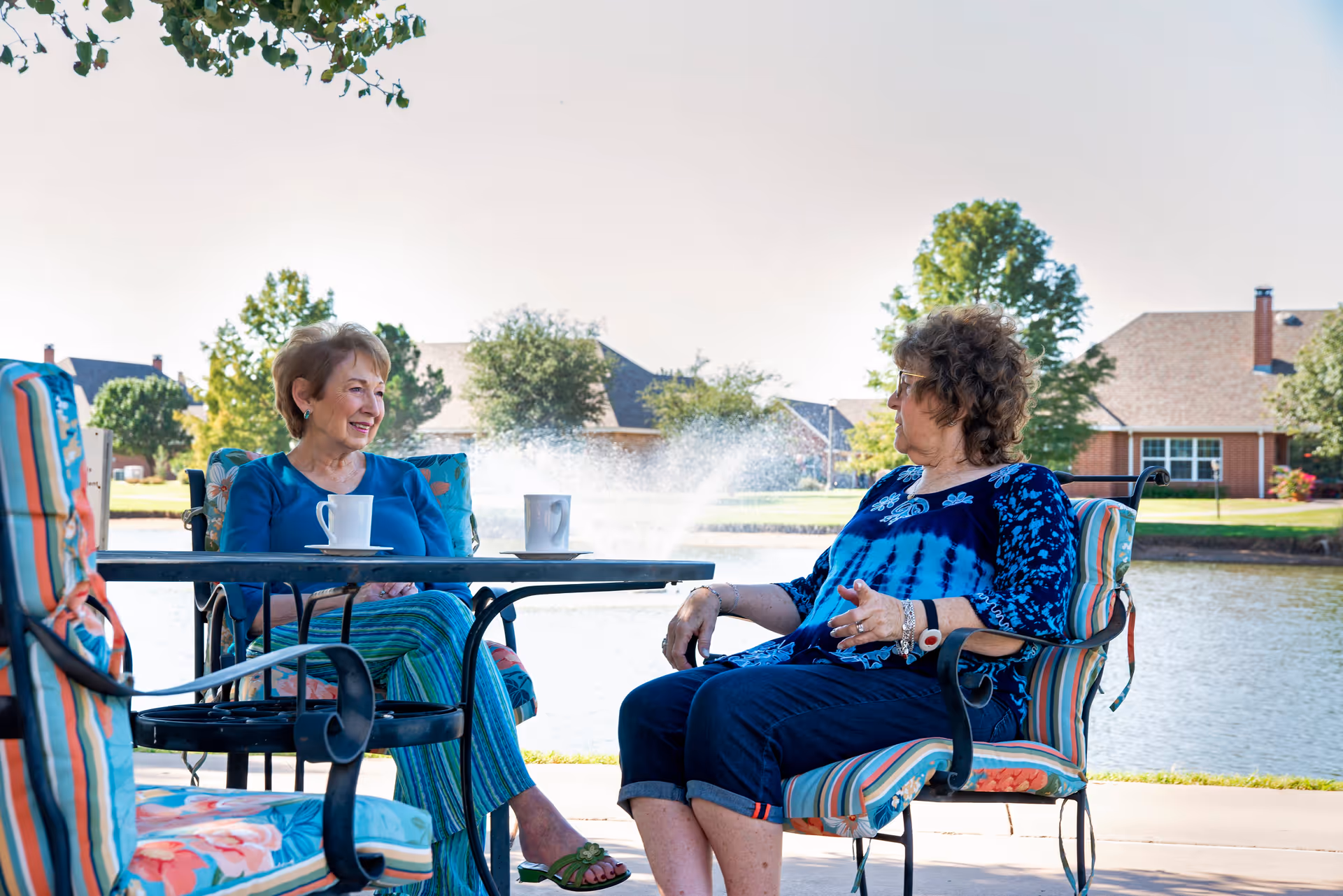 Two elderly women sitting outdoors on cushioned patio chairs around a table with coffee mugs, having a conversation by a pond with a water fountain and houses in the background.