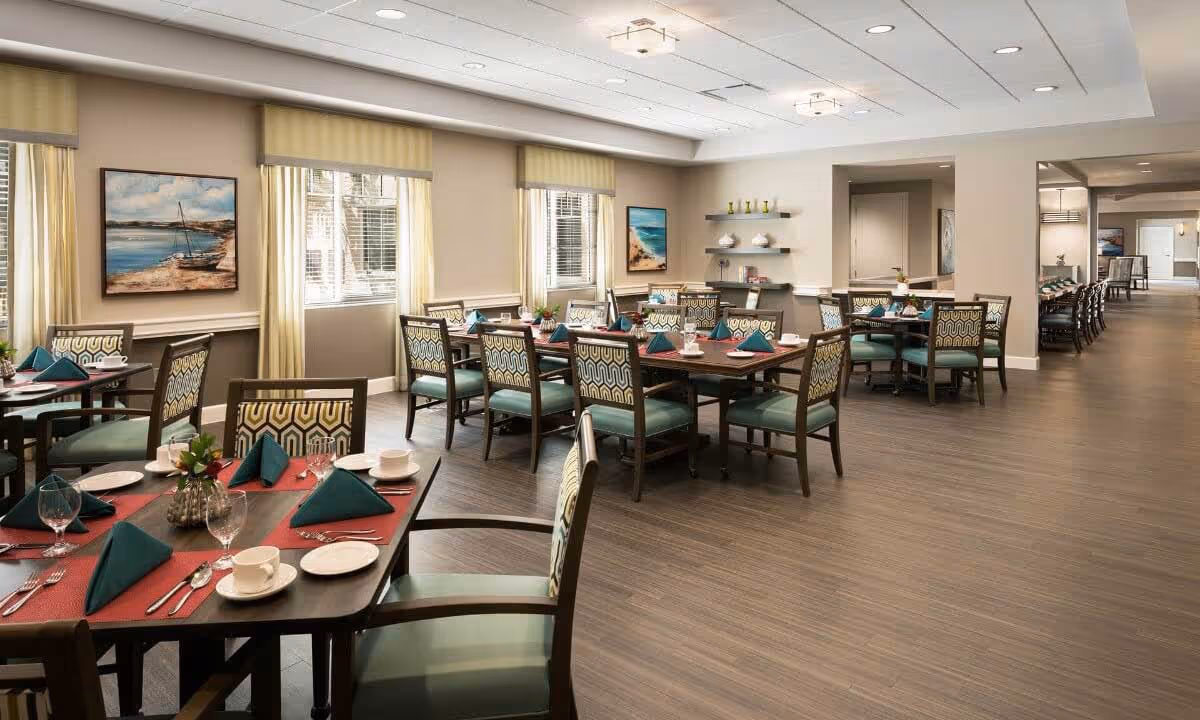 A spacious dining room in a senior living facility with multiple tables set for meals. Each table has teal napkins folded in a triangle, white plates, cups, silverware, and small floral centerpieces. The room features large windows with beige curtains, framed artwork on the walls, and shelves with decorative items. The floor is wood, and the ceiling has recessed lighting.