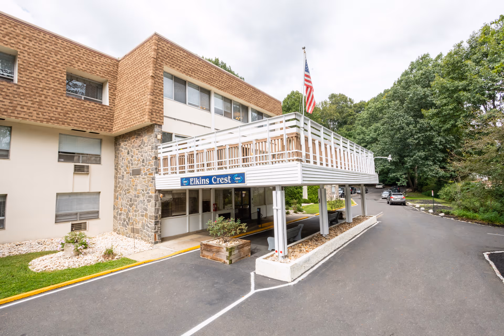 Exterior view of Elkins Crest Health & Rehabilitation Center showing the building entrance with a covered drop-off area, an American flag on a pole, and surrounding trees and parking area.