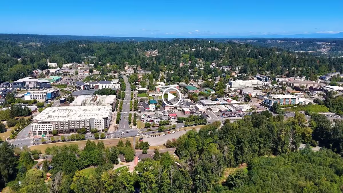 Aerial view of a suburban area with a mix of residential and commercial buildings surrounded by dense green trees and forested areas under a clear blue sky.
