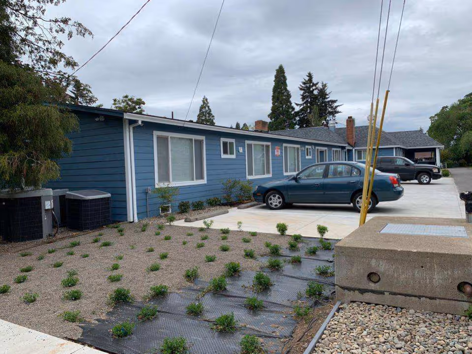 Exterior view of a single-story blue building with several windows and a parking area in front. Two cars are parked on the concrete driveway. There are small plants growing in a landscaped area covered with gravel and black weed barrier fabric. Trees and a cloudy sky are visible in the background.