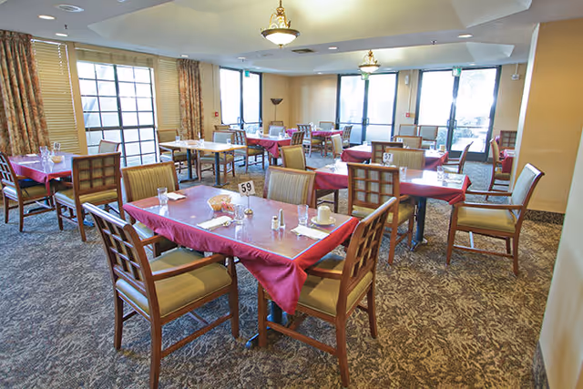 Dining room with multiple tables covered in red tablecloths, place settings, and wooden chairs beneath ceiling lights.