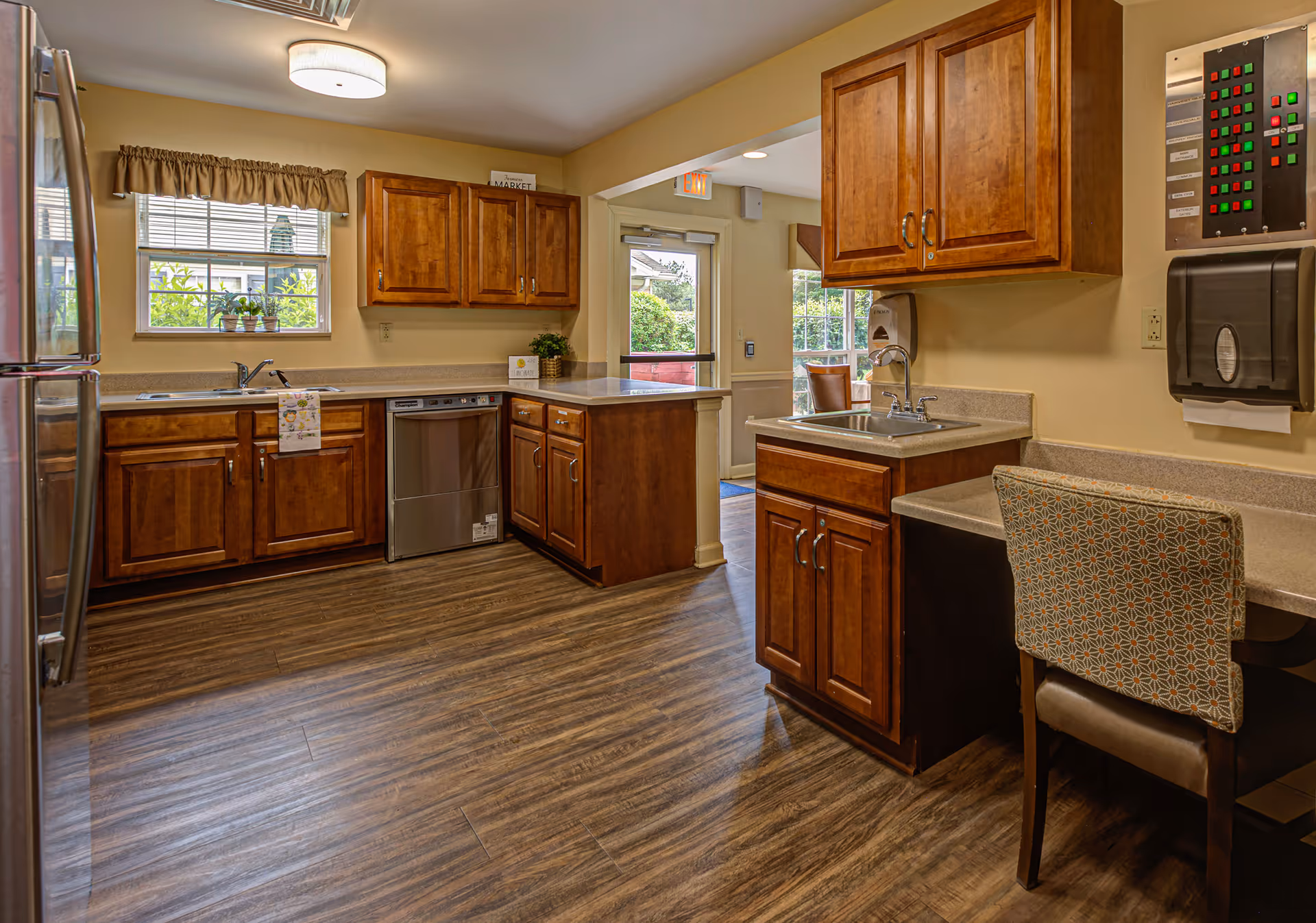 A clean and well-lit kitchen area with wooden cabinets, a stainless steel refrigerator, dishwasher, and two sinks. There is a window with a valance above one sink, a patterned chair at a small counter, and a door leading outside in the background.